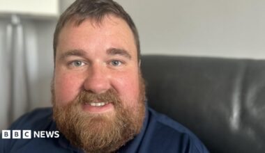 Kevin Clemitson is sitting on a black leather couch. He has mid brown hair and a lustrous beard and moustache and is fair skinned with freckles. He is smiling and wearing a navy blue top.
