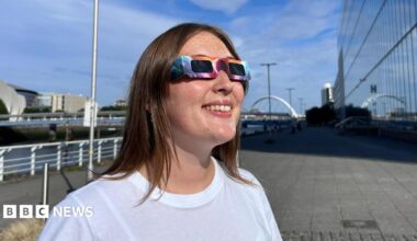 A woman wearing the eclipse glasses looking up at the sun. The glasses have a colourful pattern and black lenses. She is wearing a white T-shirt.