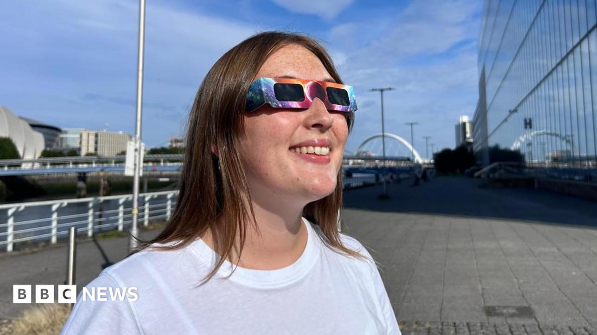 A woman wearing the eclipse glasses looking up at the sun. The glasses have a colourful pattern and black lenses. She is wearing a white T-shirt.