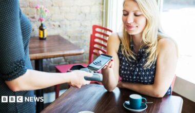 Woman sitting in a cafe with a coffee cup on the table in front of her puts her phone on a payment terminal held by a waitress.