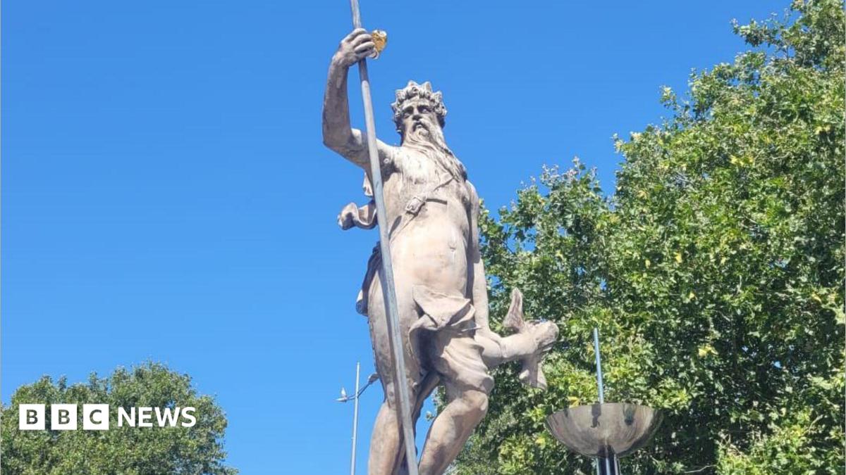 A stone sculpture of Neptune in front of green trees and a blue sky.