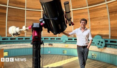 Dr Matt Bothwell standing next to a telescope. He is smiling and wearing a white shirt and black jeans. He is resting his hand on a large telescope. He is standing inside a circular wood-paneled observatory.