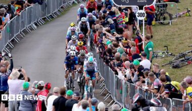 A peloton of cyclists riding on a countryside road. People are crowded around, watching from behind metal barriers.