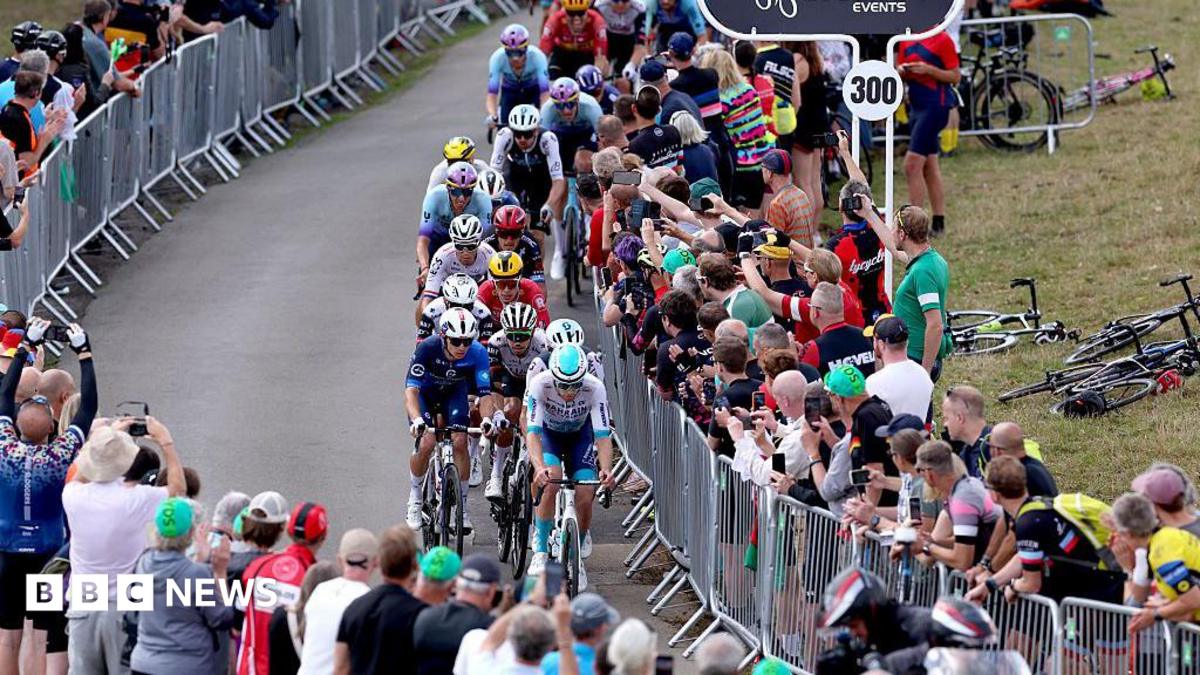 A peloton of cyclists riding on a countryside road. People are crowded around, watching from behind metal barriers.
