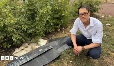 Johnny Luk wearing a white shirt and dark trousers crouches beside a small pile of discarded black plastic panels and rubble on a grassy verge next to shrubs. A paved path runs behind the bushes in an outdoor landscaped area.