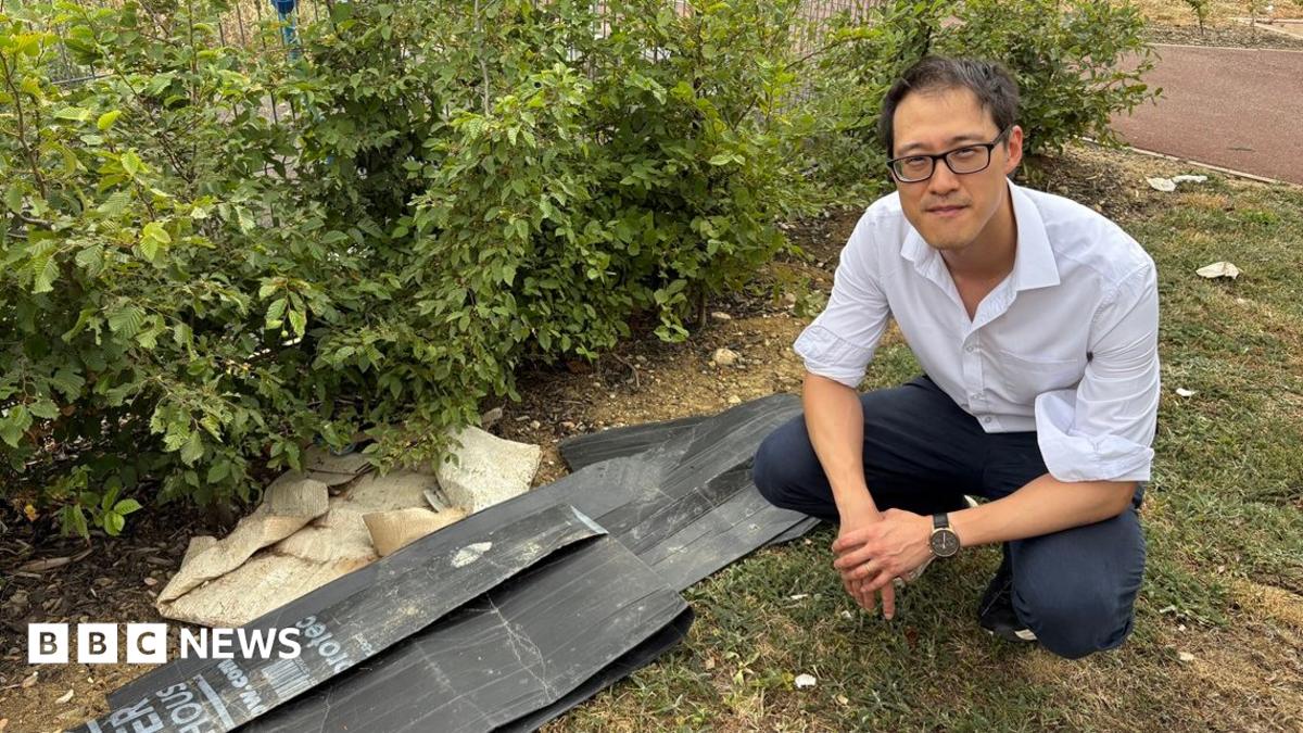 Johnny Luk wearing a white shirt and dark trousers crouches beside a small pile of discarded black plastic panels and rubble on a grassy verge next to shrubs. A paved path runs behind the bushes in an outdoor landscaped area.