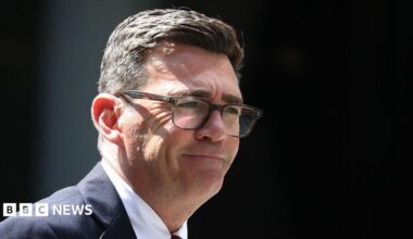 Andy Burnham is wearing glasses, a dark-coloured suit jacket, white shirt and a tie. He has a short, dark hair.