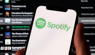 A photo of a hand holding a phone which has the green Spotify logo on it. Behind is a Spotify playlist in the background.