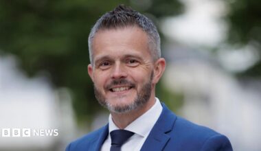 Robbie Butler. He has short grey-ish hair and beard, wearing a blue blazer, white shirt and navy tie.