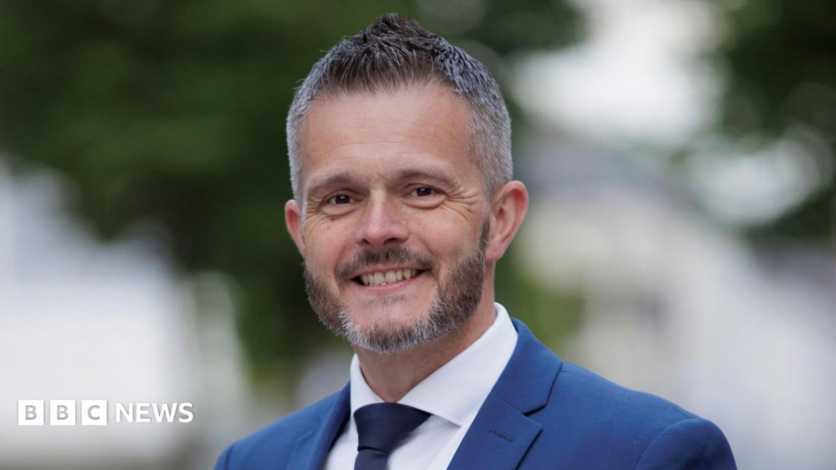 Robbie Butler. He has short grey-ish hair and beard, wearing a blue blazer, white shirt and navy tie.
