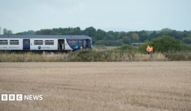 Image shows a train stopped on a railway line, with fields on either side. An official in a hi-vis jacket can be seen standing in front of it.