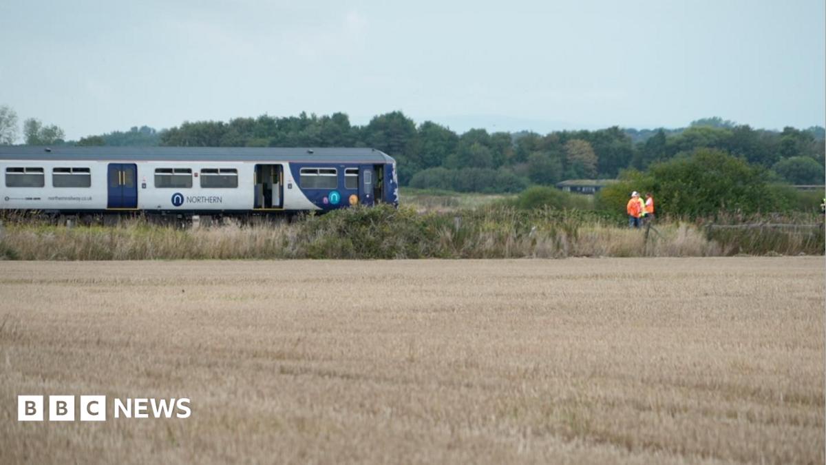Image shows a train stopped on a railway line, with fields on either side. An official in a hi-vis jacket can be seen standing in front of it.