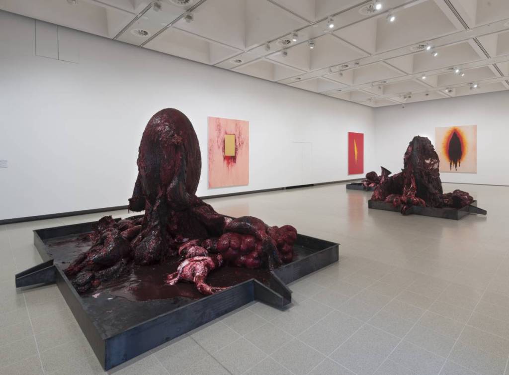 a room at the anish kapoor exhibition at the hayward gallery with paintings hung on the walls, and sculptural pieces in the middle of the room, that appear to depict horrifying gory scenes, through suggestive colours, shapes, and combinations