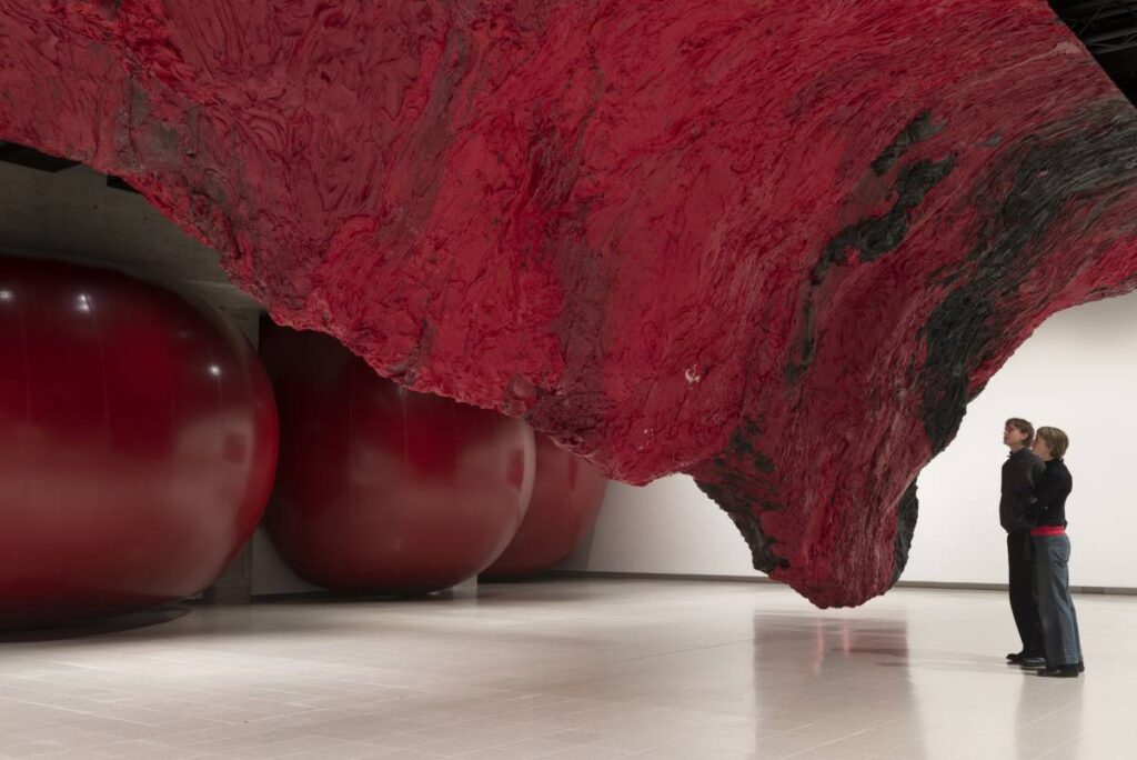 two people stood beneath an imposing large red and black scuplture that looms down from the ceiling like an upside down mountain or a strange organic lifeform