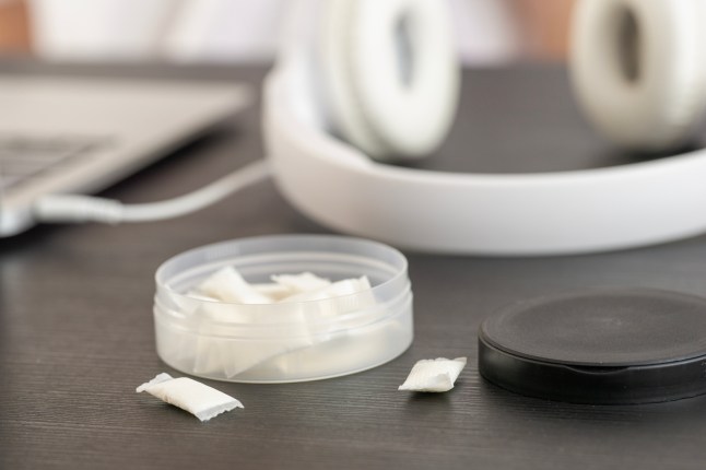 an open round plastic container with white nicotine pouches and snus pads on a dark wooden desk setup with white overhead headphones and a blurry laptop in the background. (Credits: Getty Images)