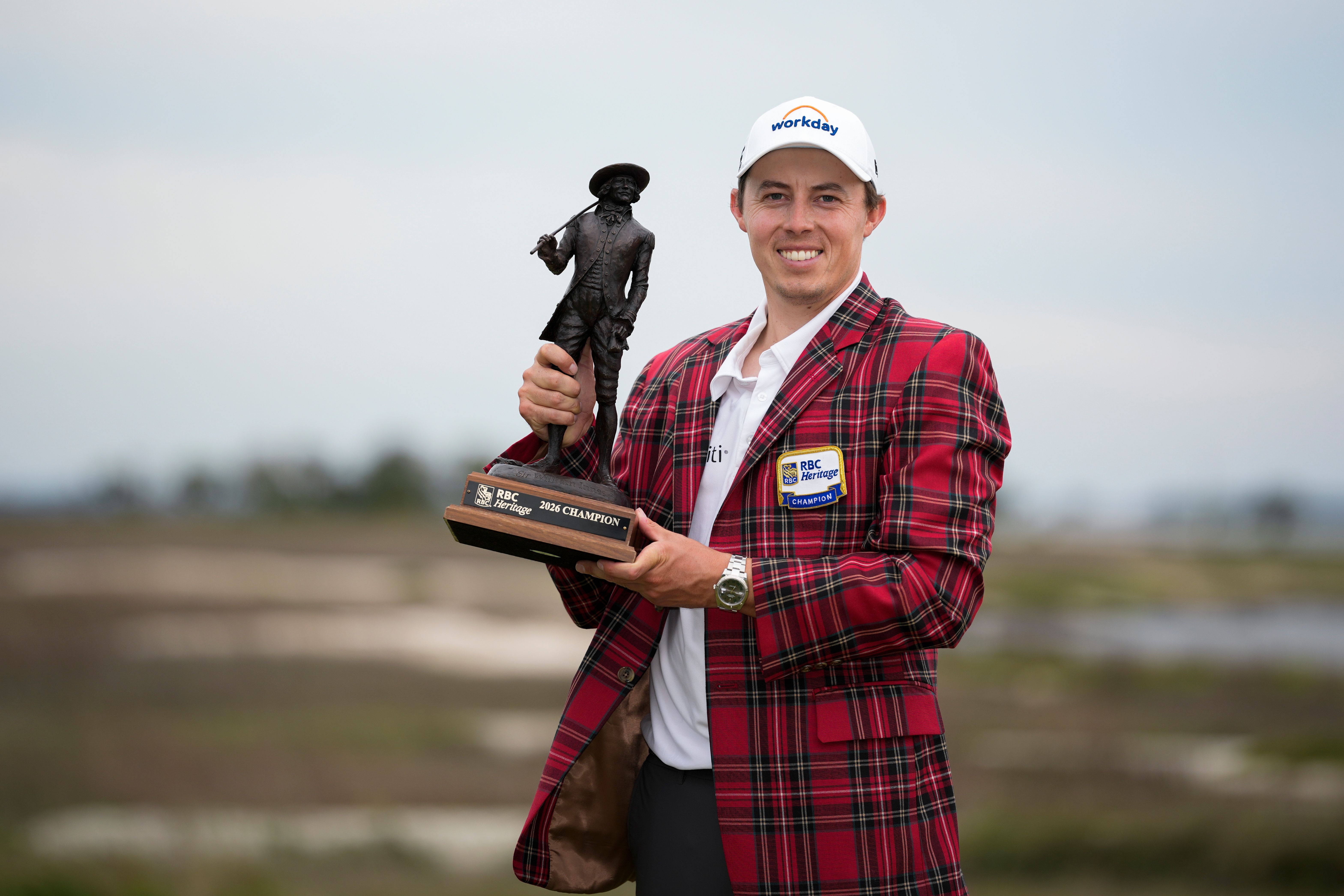 Smiling golfer in red plaid jacket and white cap proudly holds a large trophy.