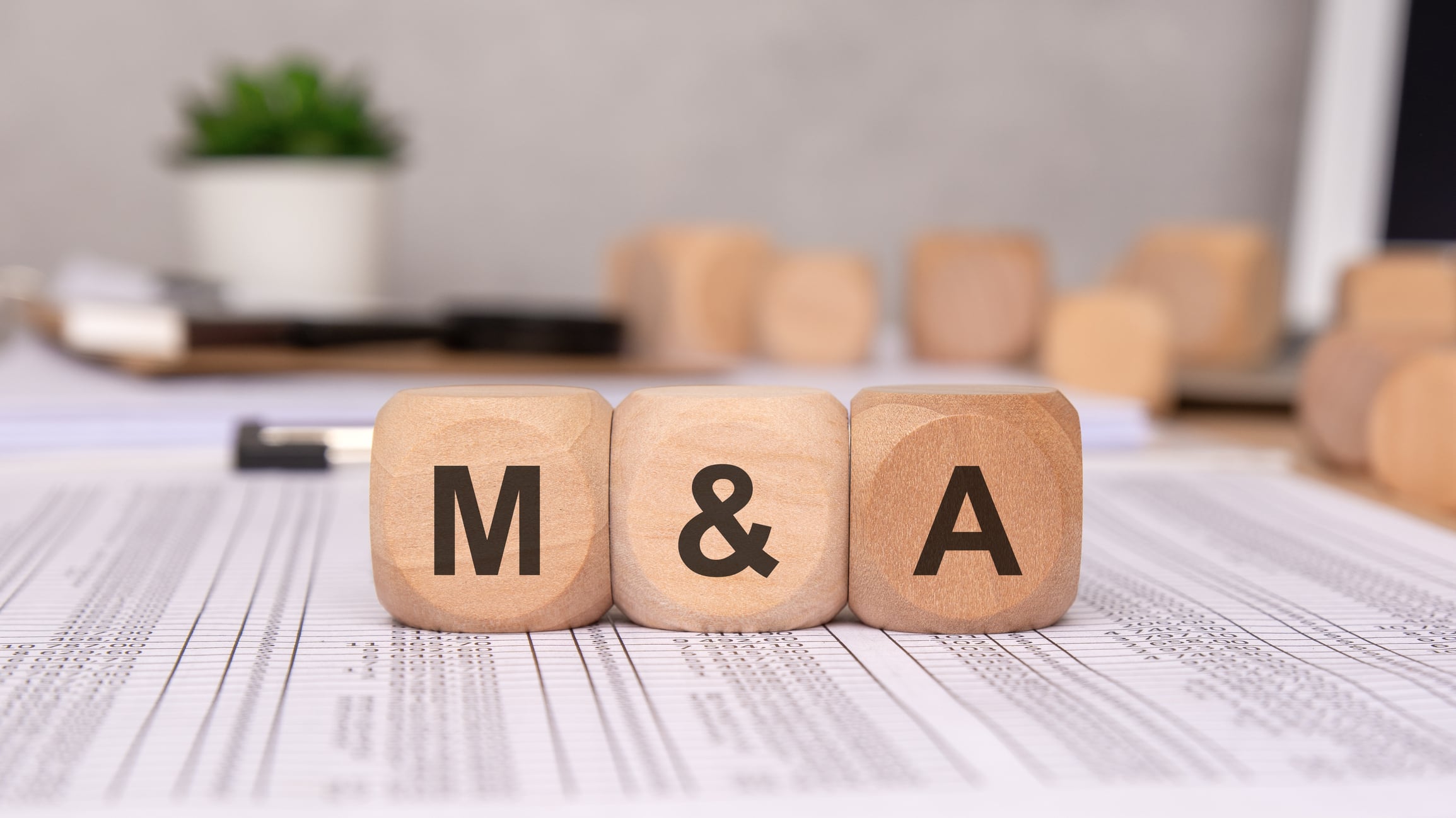 Three wooden cubes on financial spreadsheets, representing business mergers and acquisitions. blurred office background, with documents and a laptop, emphasizes corporate finance, investment strategy