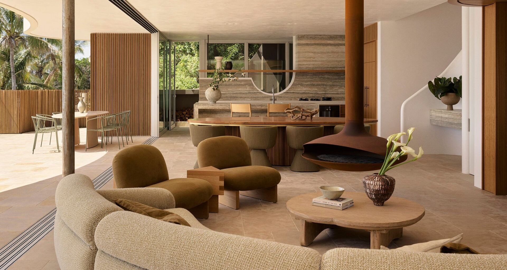 An earthy living room with a curved sectional sofa, brown suede chairs, a wood coffee table, a vase of tulips, a brown hanging fireplace, a stone kitchen, and the balcony doors drawn open
