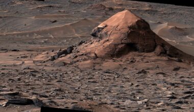A large, conical rock formation sits in a vast, rocky Martian landscape covered in scattered stones and wind-swept sand dunes.