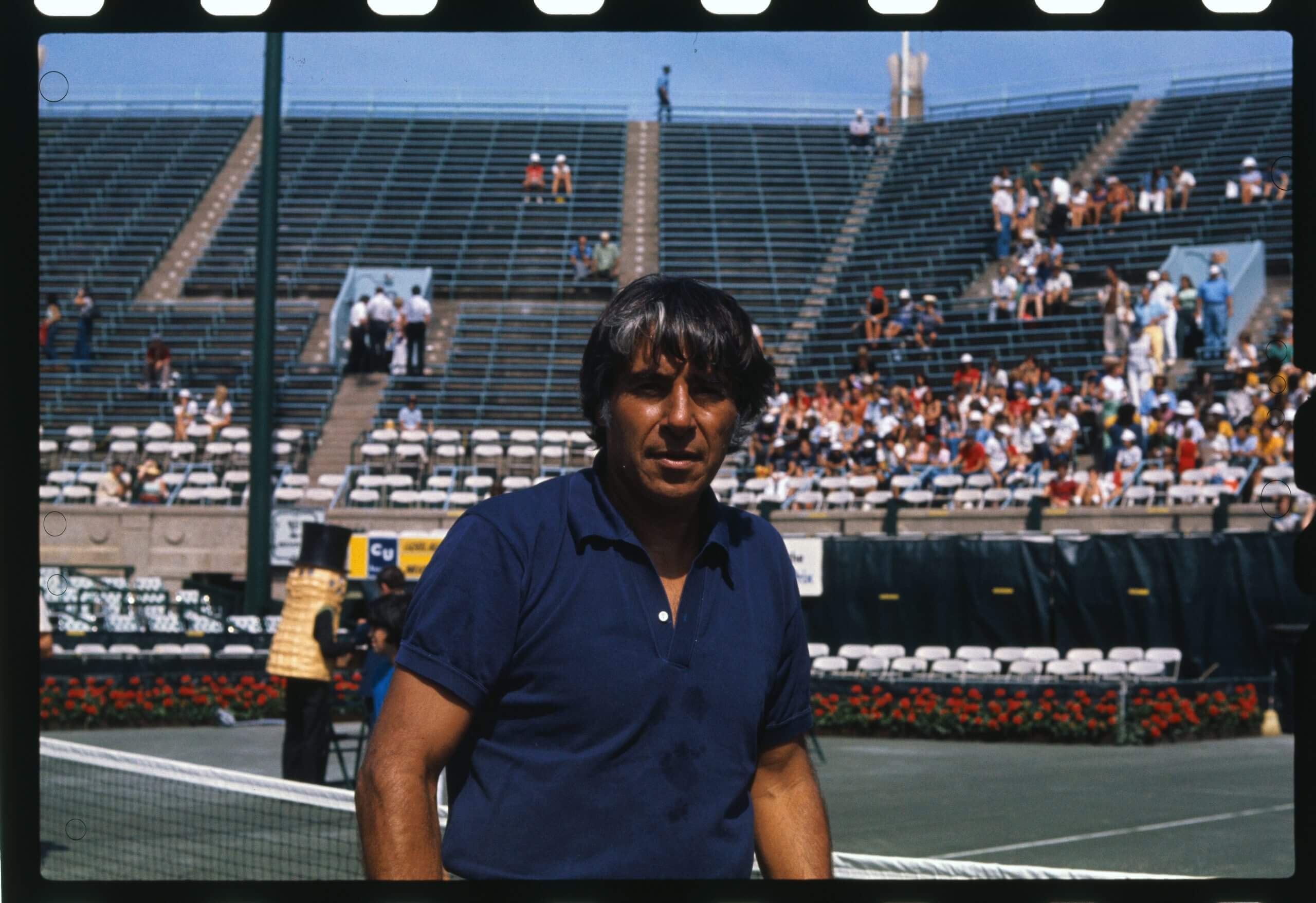 Pancho González stands in front of a tennis court with people in the crowd.