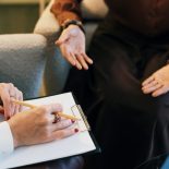 A counselor writes on a clipboard while speaking with a patient whose open hands express vulnerability and trust, conveying support, active listening, confidentiality and compassionate care in a private therapy session.