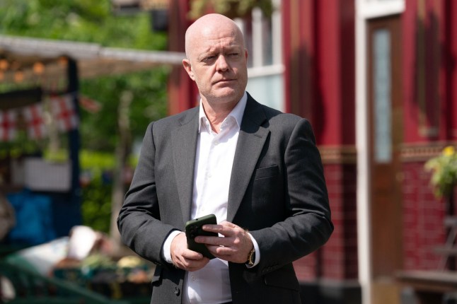 Max Branning standing in the market in EastEnders, holding his smartphone and looking thoughtful.