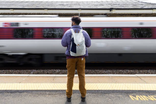 Rear-view full-length shot of a mid-adult male standing on a railway station platform in Northumberland, North East England. He is wearing a warm coat and backpack as he waits for a train to arrive. There is a blurred motion train passing by on the other side of the tracks. Videos similar to this scenario available.
