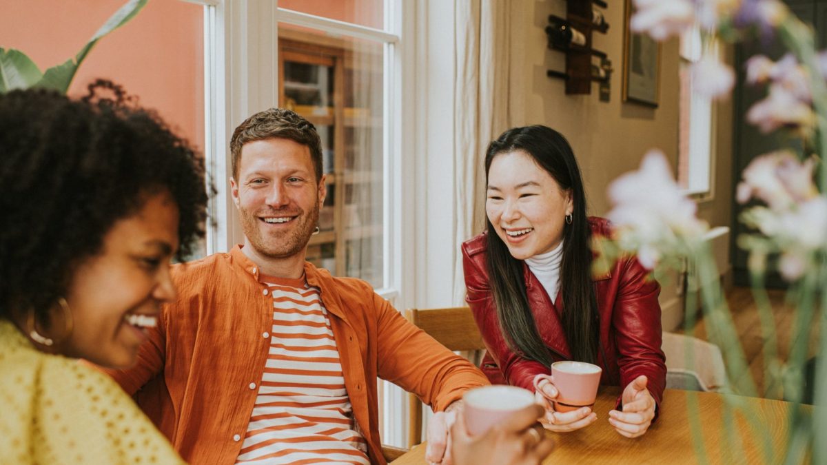 Three beautiful young people socialise at a table while enjoying hot drinks. They are animated as they giggle together. One girl is in the foreground whilst focus remains on a smiling male and female.
