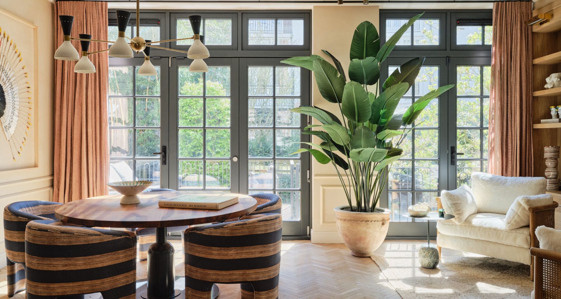 Dining area with brown and blue striped tub chairs, round pedestal table, black Crittall-style doors, apricot curtains, white armchairs, large potted plant and multi-light pendant
