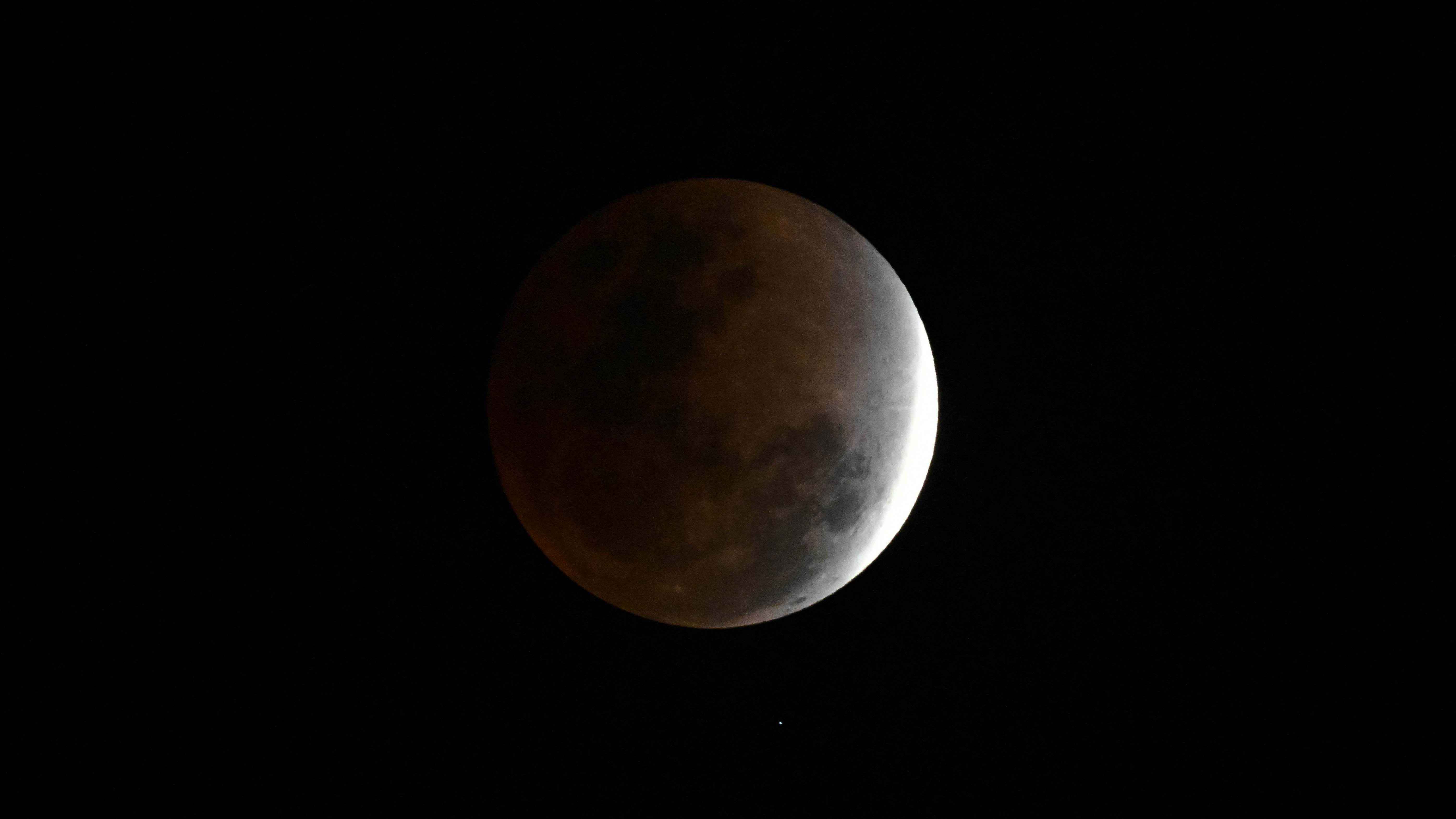 The moon is photographed in a black sky during a total lunar eclipse with a thin crescent on its right side exposed to direct sunlight. Dark lunar seas are visible on its red-tinged shadowed side.