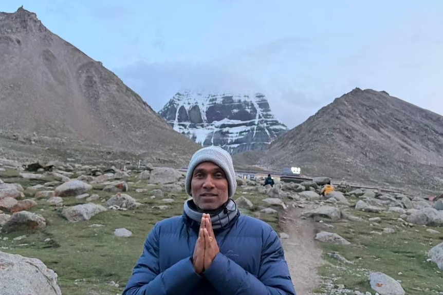 A man in a beanie and puffer jacket holds his hands as if in prayer while standing on a track in front of a mountain range.