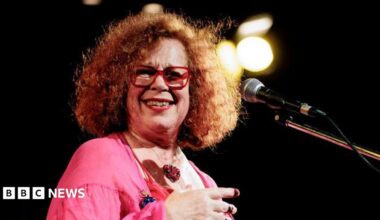 A woman, Sarah Jane Morris, wearing a pink top and standing on a stage with a microphone.