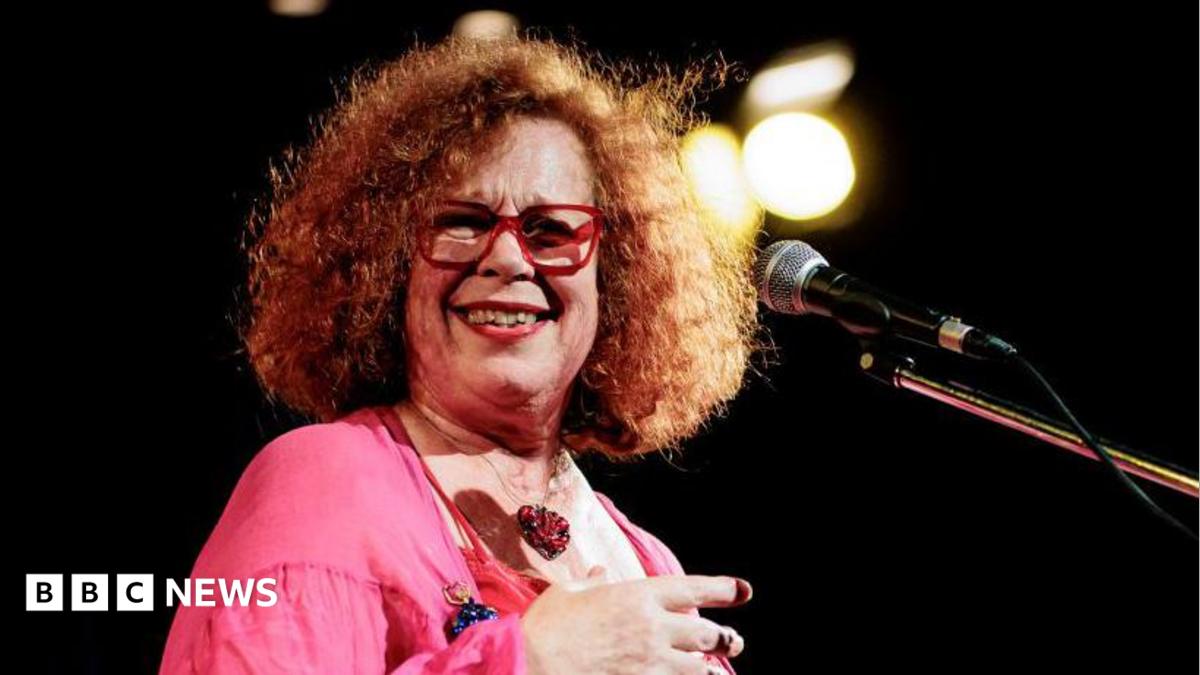 A woman, Sarah Jane Morris, wearing a pink top and standing on a stage with a microphone.