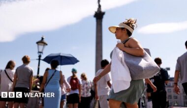 A woman with a sunhat and visor on walks through Trafalgar Square with other people holding umbrellas in the background to shield them from the sun. With Nelson's Column in the background