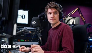 Greg James wears a burgundy jumper. He is sitting in front of a radio desk with headphones on.