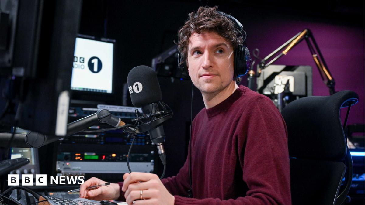 Greg James wears a burgundy jumper. He is sitting in front of a radio desk with headphones on.