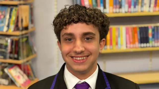 Laith has short curly brown hair and smiles at the camera. He wears a black school blazer, white shirt and purple tie and appears to be in a library