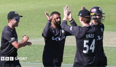 Ryan Patel raises his bat to the crowd after scoring a half-century. He is wearing an all black Surrey One-Day Cup kit
