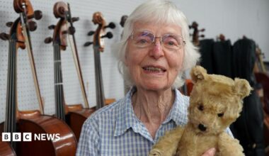 Jaquelin du Pre's sister smiles at the camera and holds an old teddy bear in front of a row of cellos