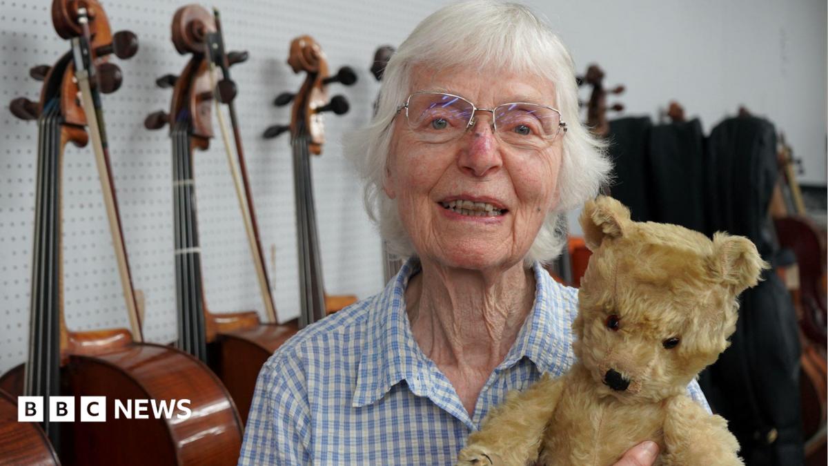 Jaquelin du Pre's sister smiles at the camera and holds an old teddy bear in front of a row of cellos