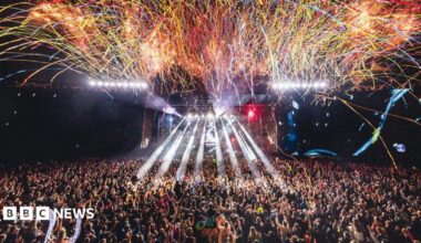 A large crowd of people are watching a performance at Creamfields as streamers fall from above and rays of white light are projected from the stage.