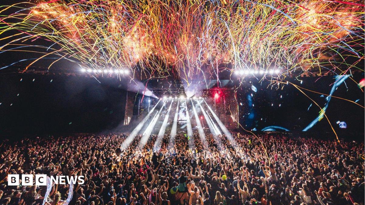 A large crowd of people are watching a performance at Creamfields as streamers fall from above and rays of white light are projected from the stage.