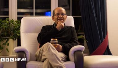 An elderly man sits in a chair and smiles. He looks like he might be speaking. He holds a class in his lap and sits upright in a navy sweater.