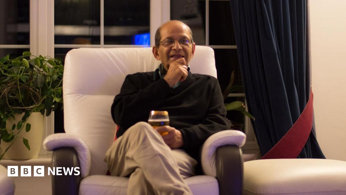 An elderly man sits in a chair and smiles. He looks like he might be speaking. He holds a class in his lap and sits upright in a navy sweater.