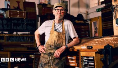 Nick Hand wearing a grey t-shirt and brown apron, which is heavily stained with black ink. He is leaning on a wooden table inside his workshop, looking straight at the camera with his hand on his hip.