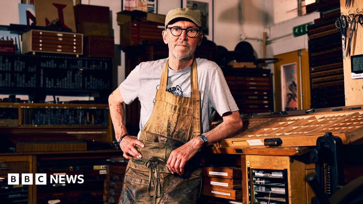 Nick Hand wearing a grey t-shirt and brown apron, which is heavily stained with black ink. He is leaning on a wooden table inside his workshop, looking straight at the camera with his hand on his hip.