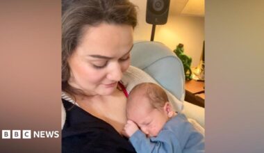 A mother is holding her son who is a newborn. The baby is lying into her chest and is asleep. She is looking at him tenderly.
