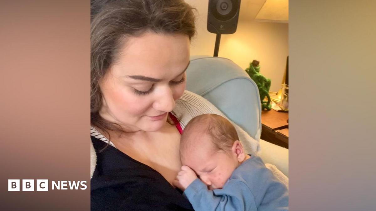 A mother is holding her son who is a newborn. The baby is lying into her chest and is asleep. She is looking at him tenderly.