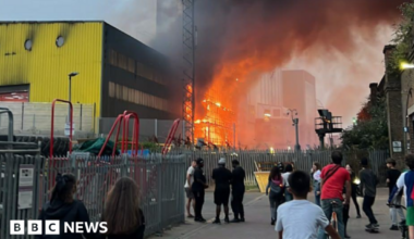 Bright orange flames and thick black smoke engulf a structure behind a metal fence and a telecommunications tower, next to a large yellow and black industrial building under a hazy dusk sky.