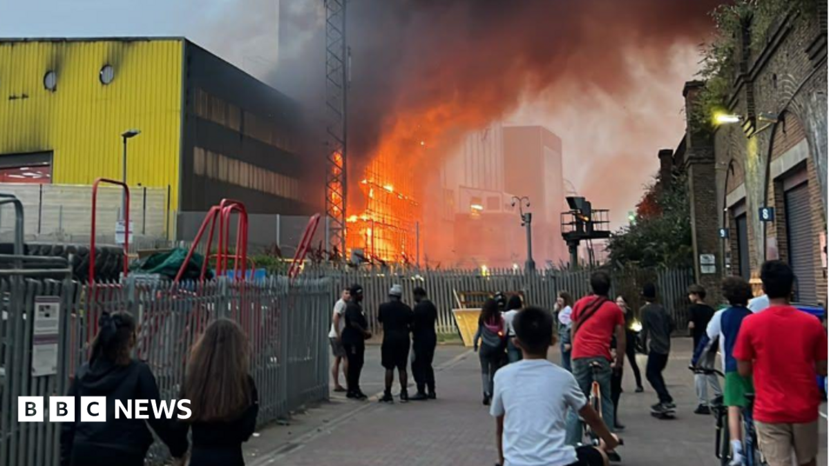 Bright orange flames and thick black smoke engulf a structure behind a metal fence and a telecommunications tower, next to a large yellow and black industrial building under a hazy dusk sky.